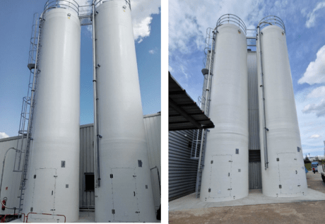 Installation of two white industrial silos side by side, equipped with ladders, walkways, and piping, in front of a building under a blue sky.