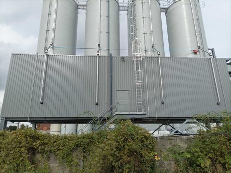 Four white industrial silos lined up on a metal platform with a safety ladder and pipes, in front of a gray panel facade.