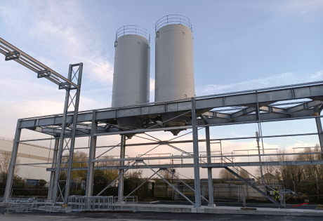 Two white industrial silos installed on an external metal frame with walkway and access structure, photographed at dusk on a production site.