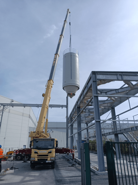 Mobile crane positioning a white industrial silo on an external metal frame at a production site.