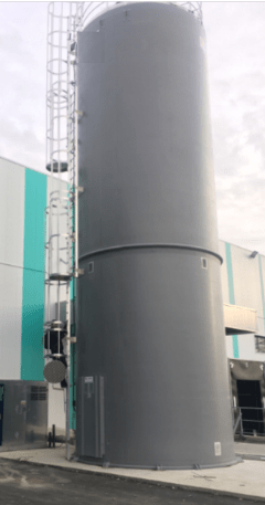 Rigid industrial silo in anthracite gray equipped with a ladder and steel guardrail, installed in front of a facade with white and turquoise panels.