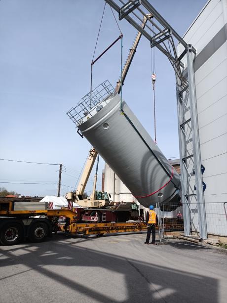 Worker wearing a high-visibility vest guiding the installation of a rigid industrial silo being tilted by a mobile crane mounted on a flatbed truck at a production site.