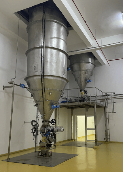 Two rigid stainless steel silos installed indoors on a metal platform with valves and piping for feeding and regulating an industrial process.