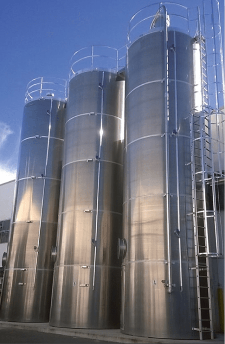 Three rigid industrial silos made of shiny stainless steel, equipped with ladders and guardrails, installed outdoors under a blue sky.